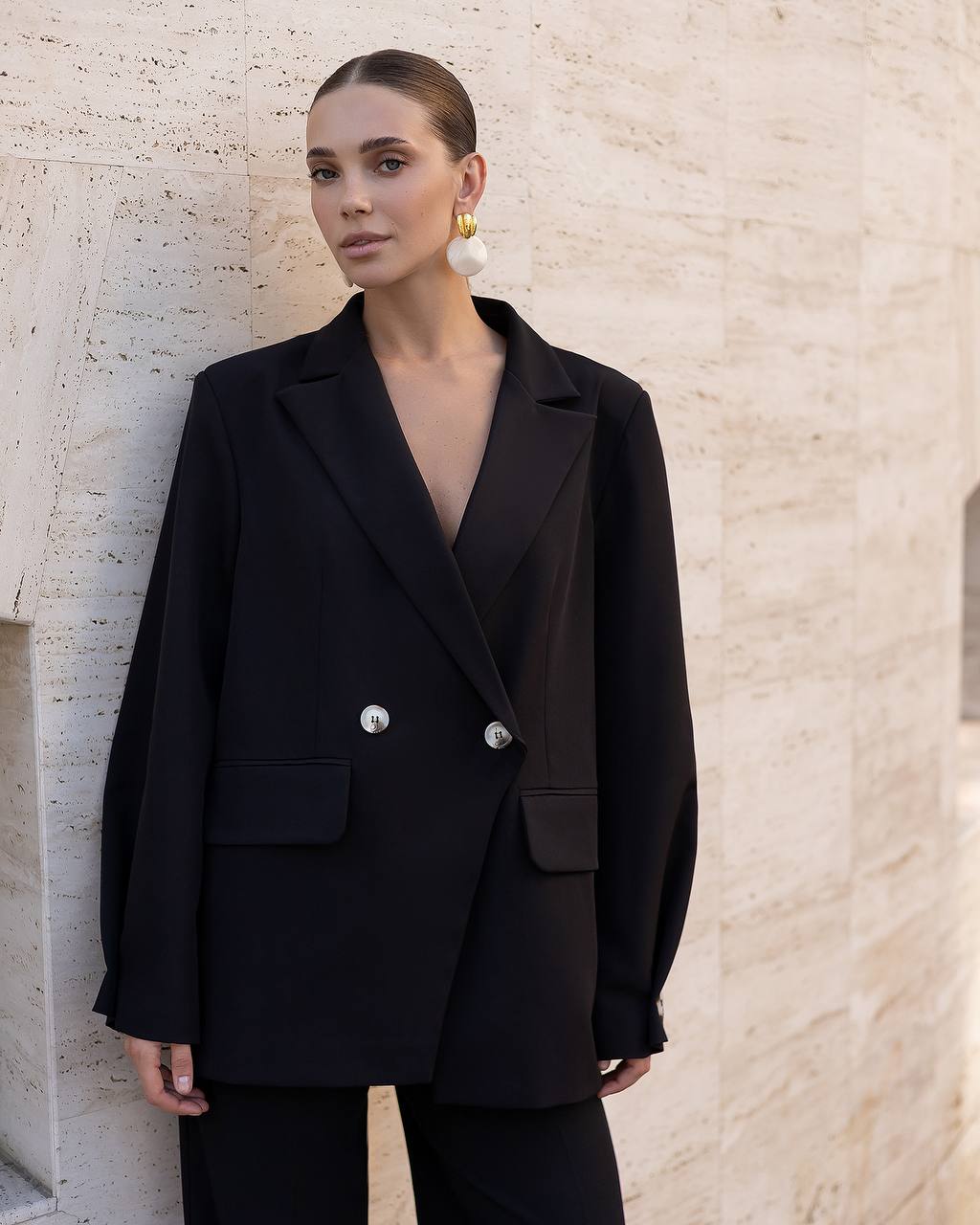 A woman wearing an oversized black double-breasted blazer with silver buttons and matching black trousers, standing against a light beige textured stone wall.