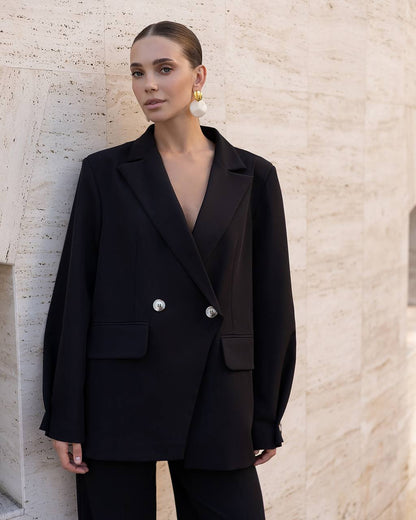 A woman wearing an oversized black double-breasted blazer with silver buttons and matching black trousers, standing against a light beige textured stone wall.