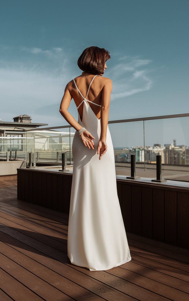 Woman in a white evening gown with a backless design on a rooftop with cityscape view.