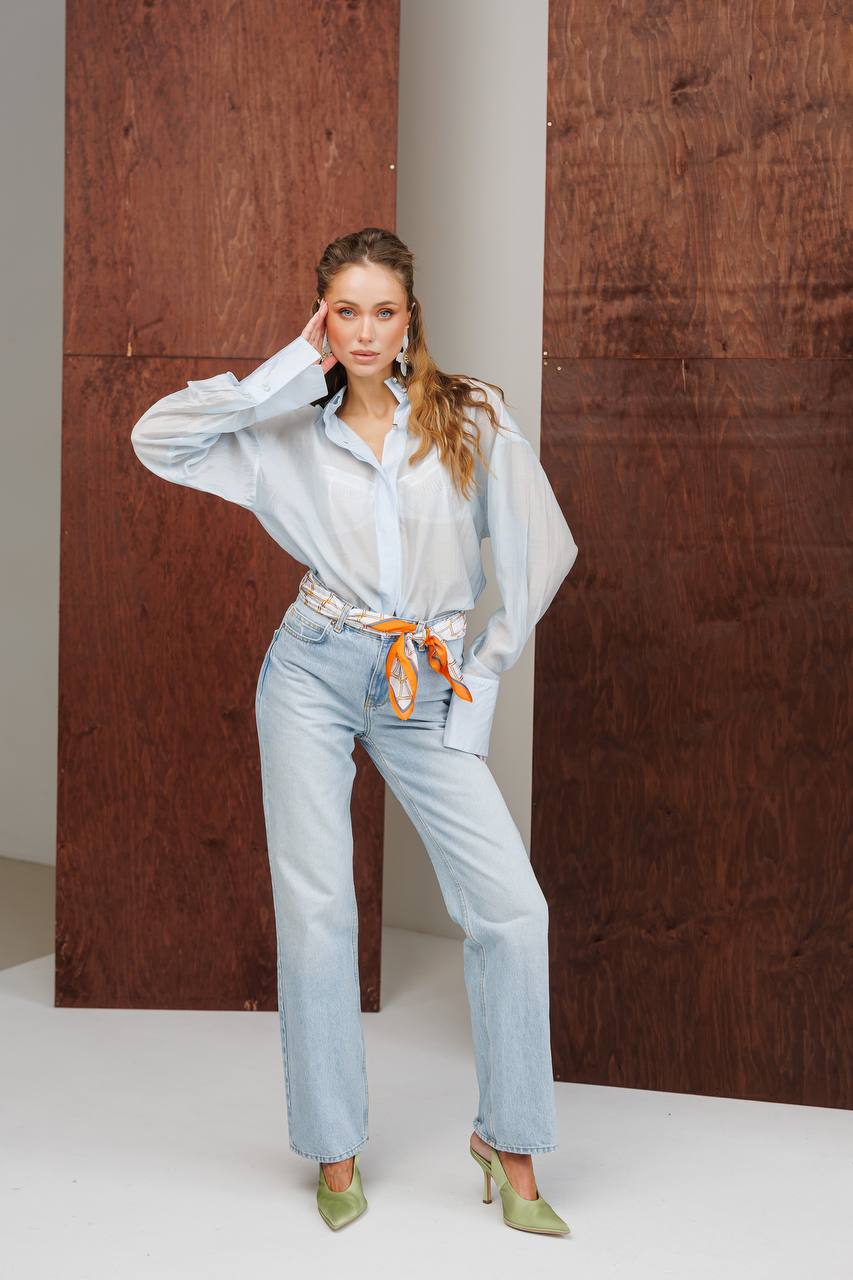 Woman wearing a light blue shirt and jeans with an orange belt, standing against a wooden wall.