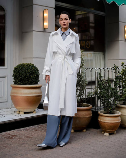 Woman in a white trench coat and blue pants standing in front of a building with plants.