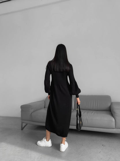Person wearing a black dress standing in front of a gray sofa against a plain wall.