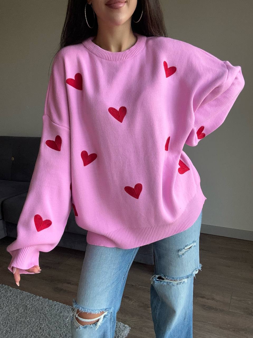 Person wearing a pink sweater with red heart patterns and ripped jeans indoors.