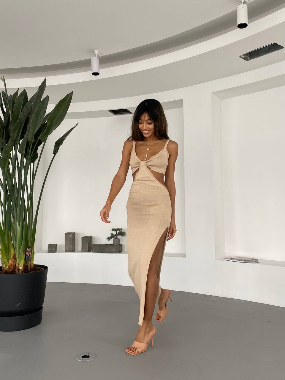 Woman in a beige dress standing in a modern indoor setting with a plant.