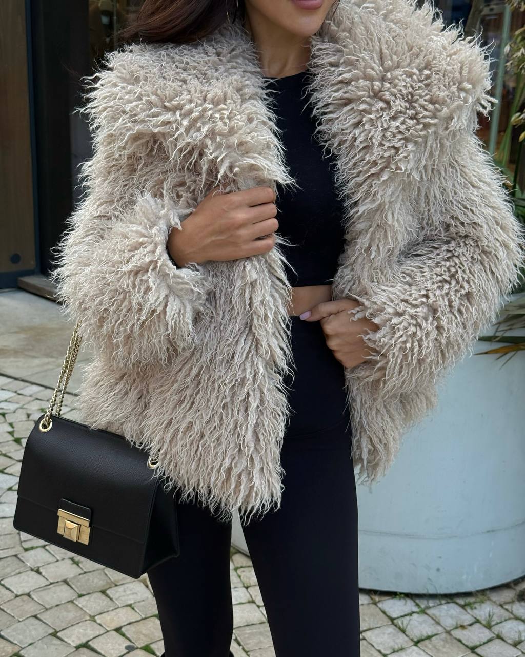 Person wearing a fluffy beige jacket holding a black handbag.