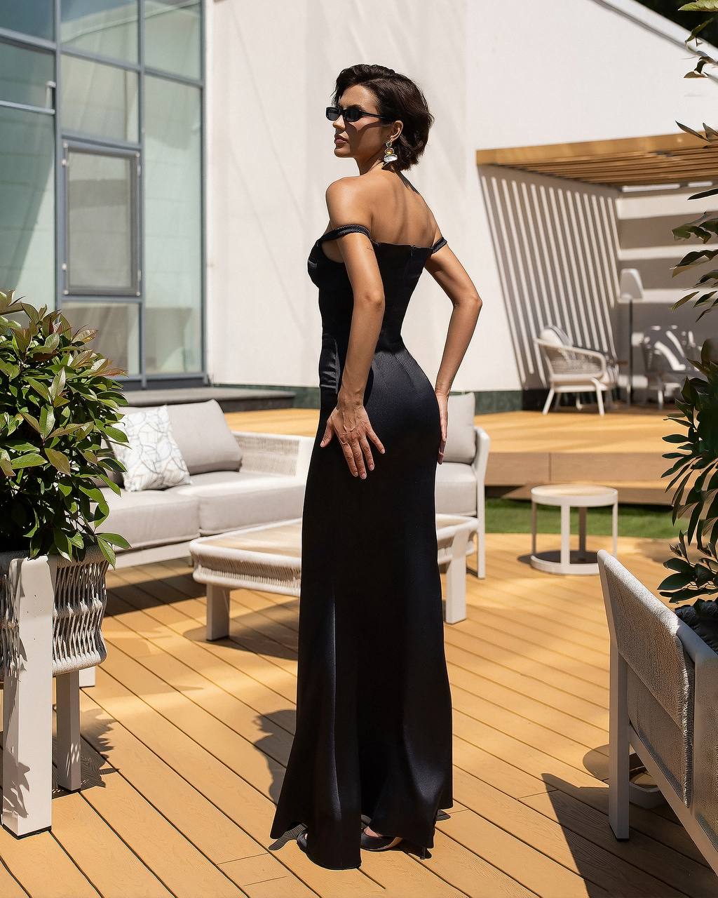 A woman wearing a floor-length black off-the-shoulder gown stands on a wooden outdoor patio, photographed from the back and side. The setting includes white patio furniture, plants, and a modern building in the background.