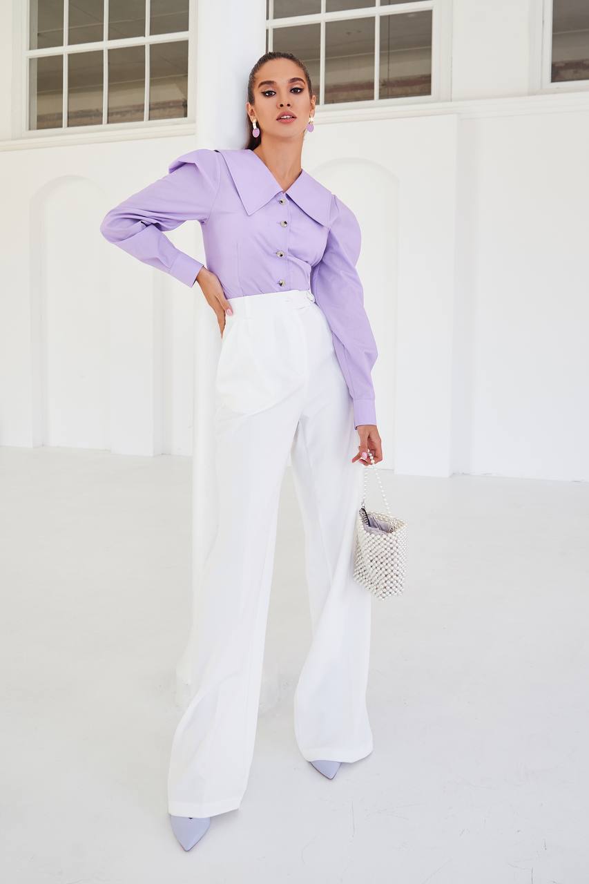 Woman wearing a light purple top and white pants standing against a white wall.