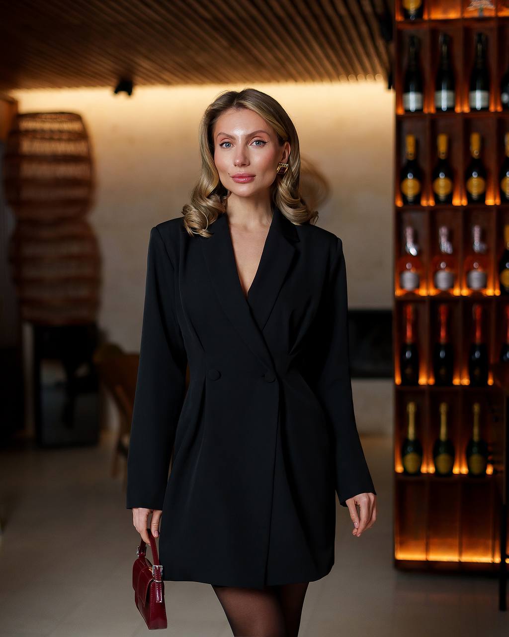 Woman in a black coat standing in a dimly lit room with wine bottles in the background