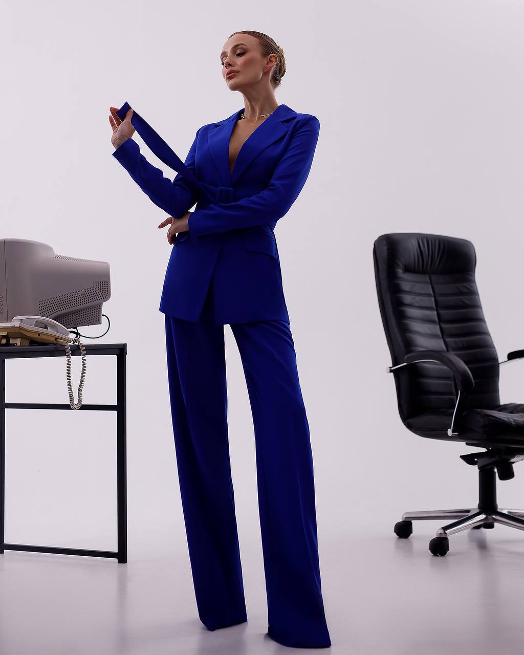 Woman in a blue suit standing in an office setting with a chair and desk.