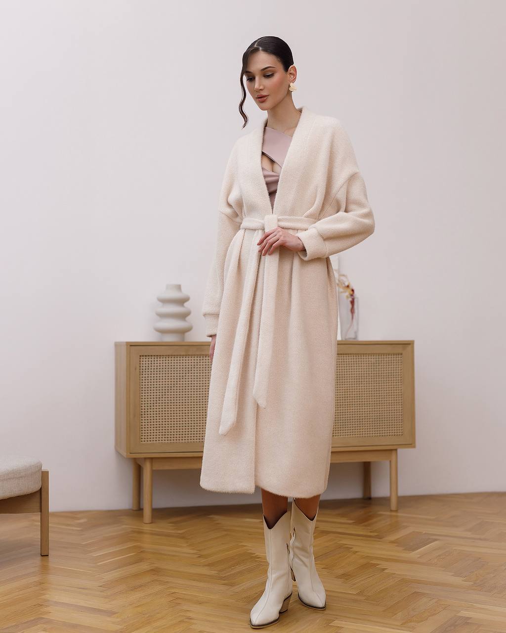 Woman wearing a long beige coat in an indoor setting with wooden flooring and a light-colored wall.