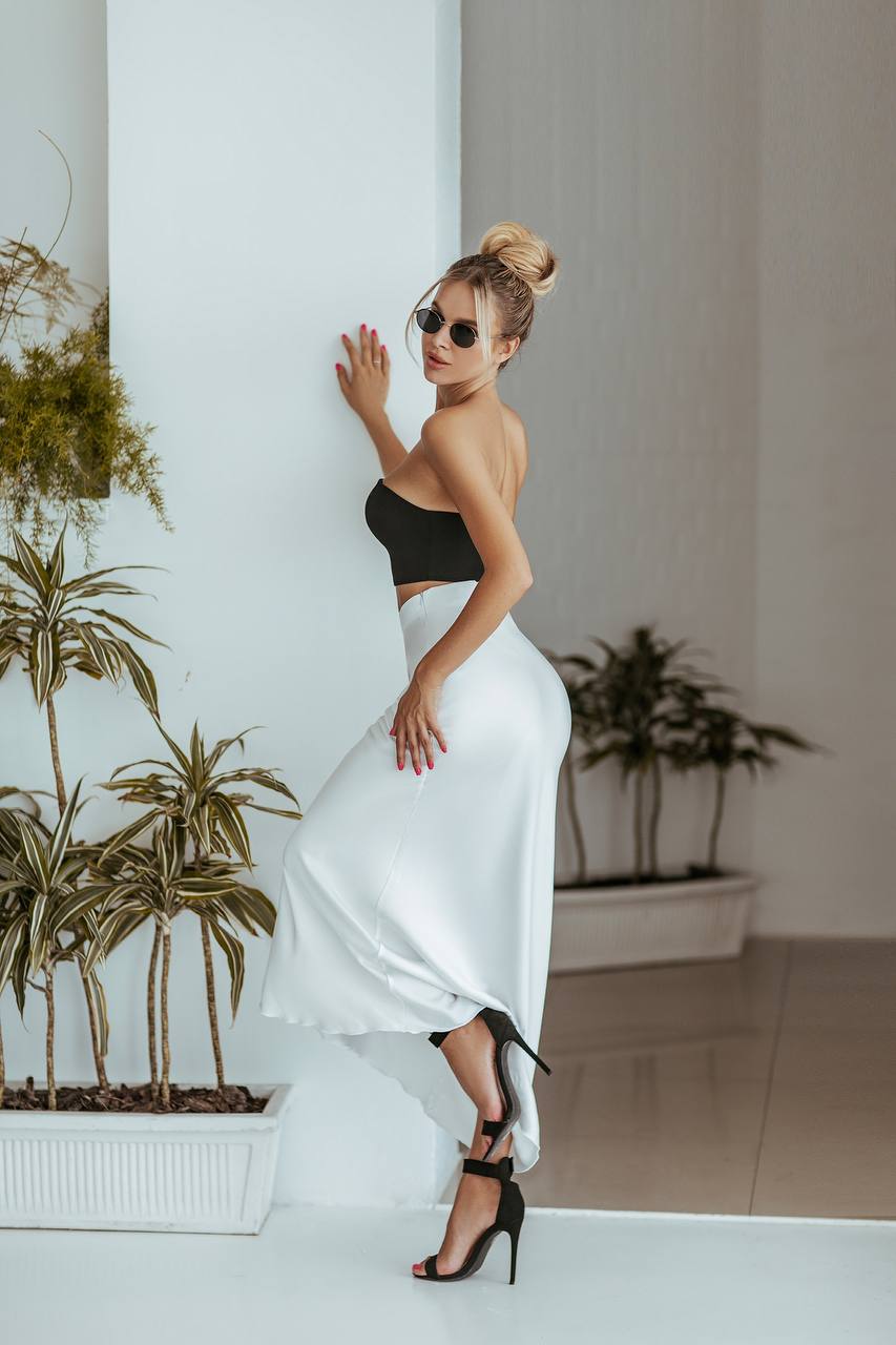 Woman in a black top and white skirt standing indoors with plants in the background