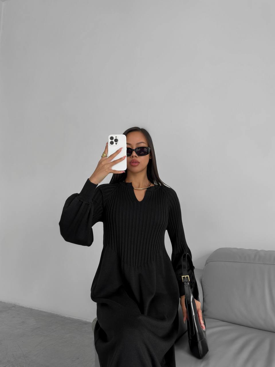 Person wearing a black outfit taking a mirror selfie against a gray wall.