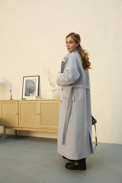 Woman wearing a long light gray coat in a room with a wooden cabinet and decorative items.