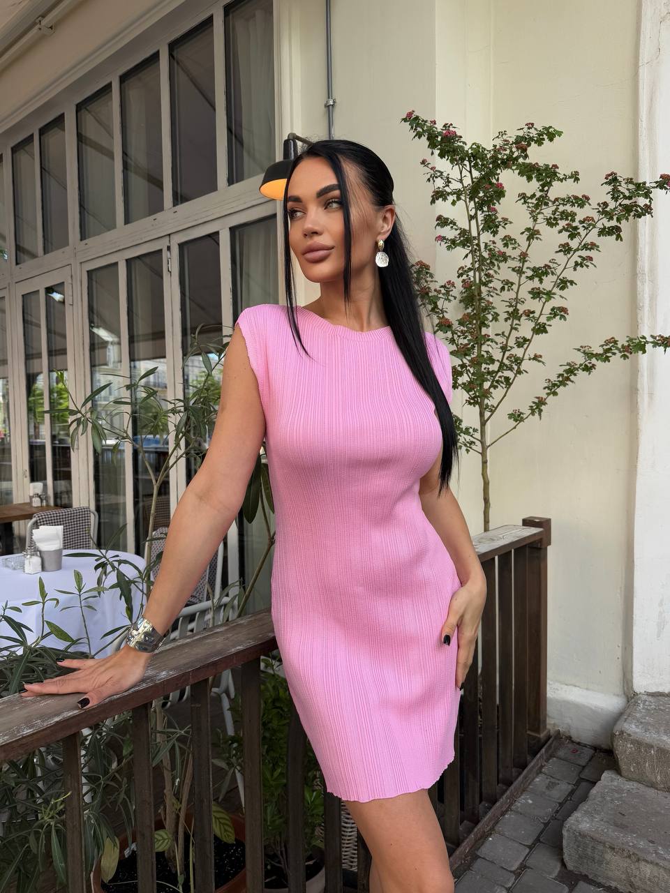 Woman in a pink dress standing outdoors near a building with plants.
