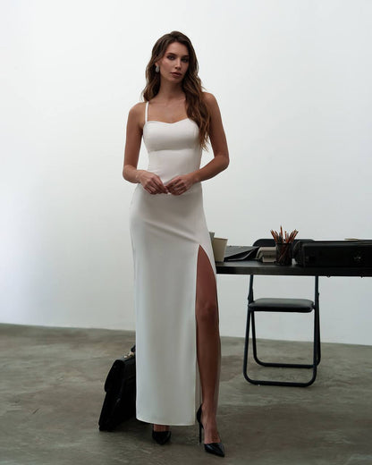 Woman in a white dress standing in an indoor setting with a table and chair.