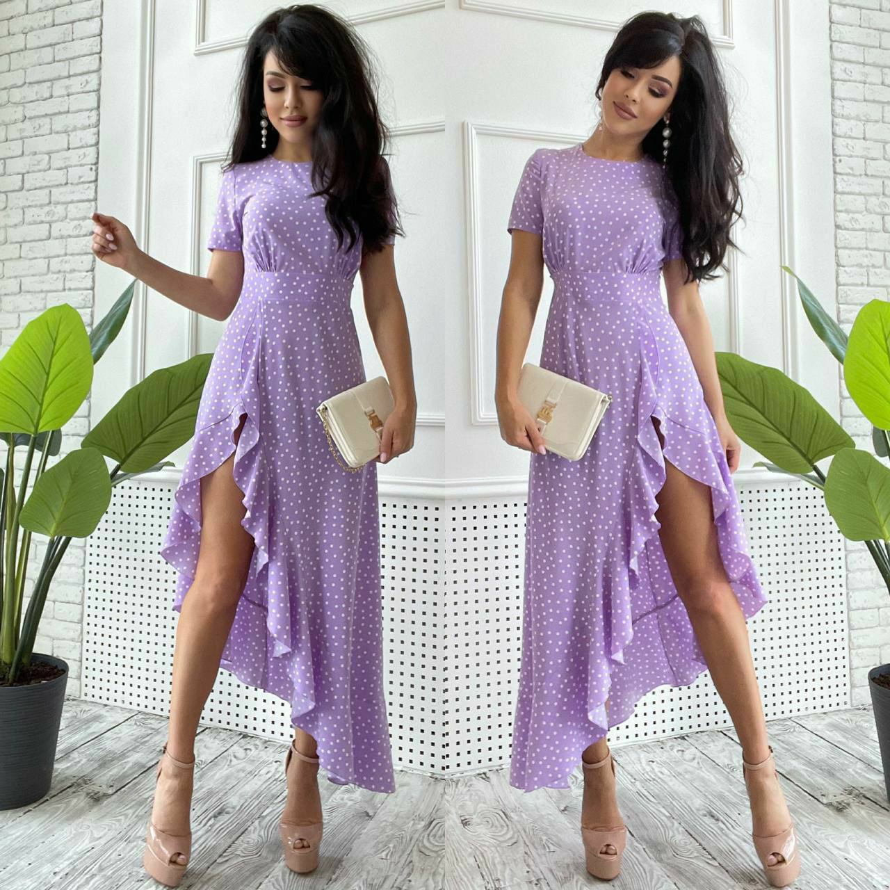 Woman wearing a purple polka dot dress with a slit, standing indoors with plants in the background.