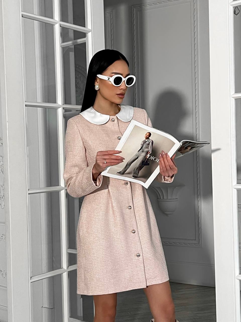 Woman in a beige dress holding a Dior magazine in a room with white walls and a door.