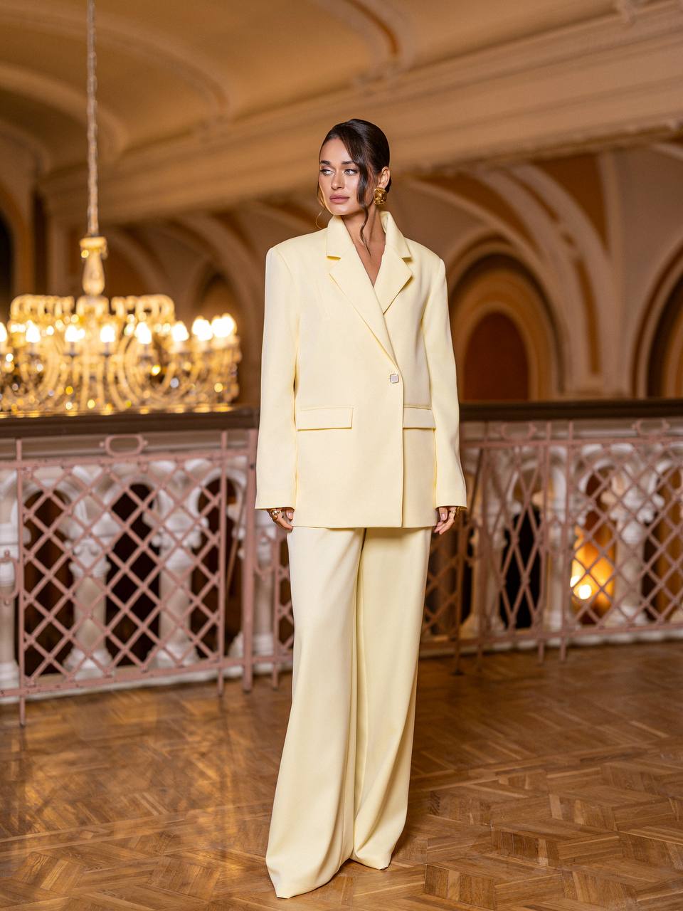 Woman in a light yellow suit standing in an elegant indoor setting with chandelier.