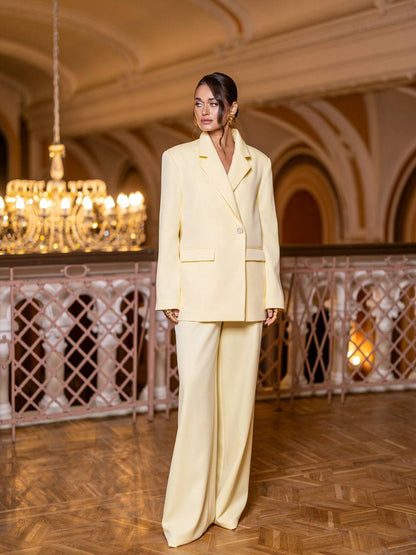 Woman in a light yellow suit standing in an elegant indoor setting with chandelier.