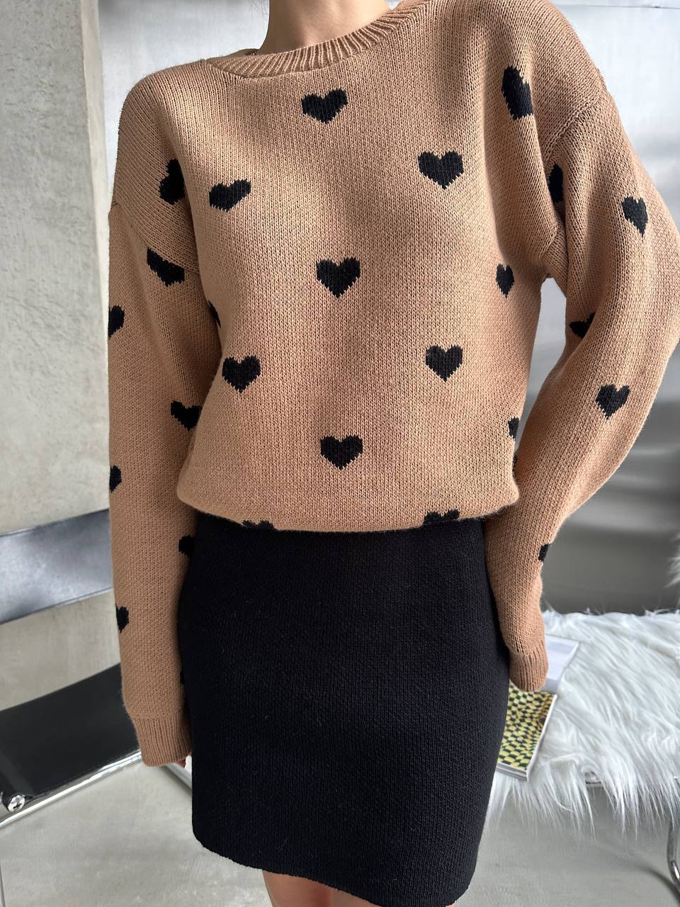 Beige sweater with black heart patterns worn by a person, standing against a neutral background.