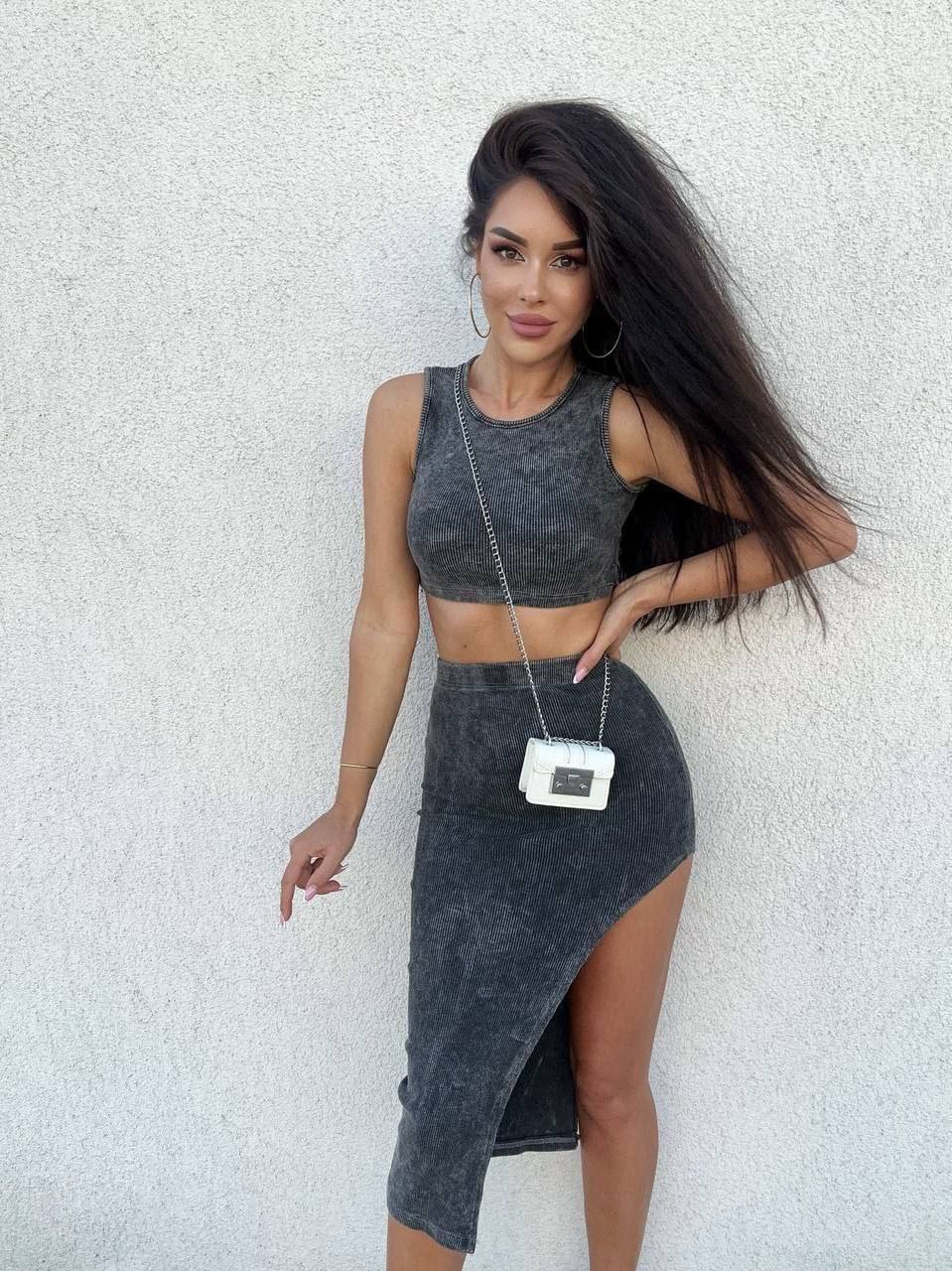 Woman in gray sleeveless top and skirt with a white handbag against a light gray wall.