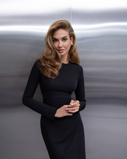 A woman with blonde hair wearing a black long-sleeve dress, standing against a gray metallic background.