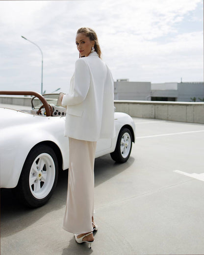 Woman in a white outfit standing next to a white car on a concrete surface with a cloudy sky.