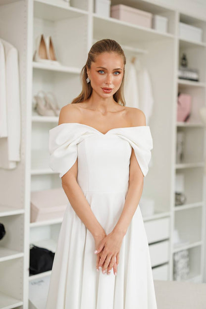 Woman wearing a white off-shoulder dress in a room with shelves.