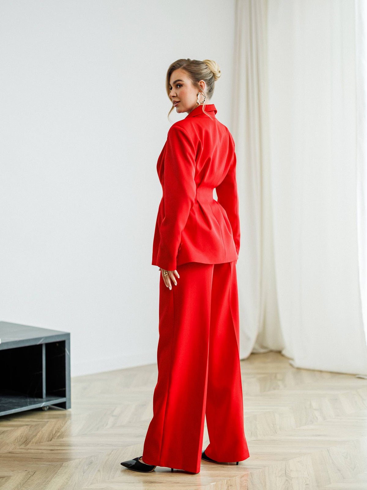 A woman wearing a bright red two-piece suit with a structured blazer and wide-leg trousers, standing in a minimalist room with light wood floors and white curtains.