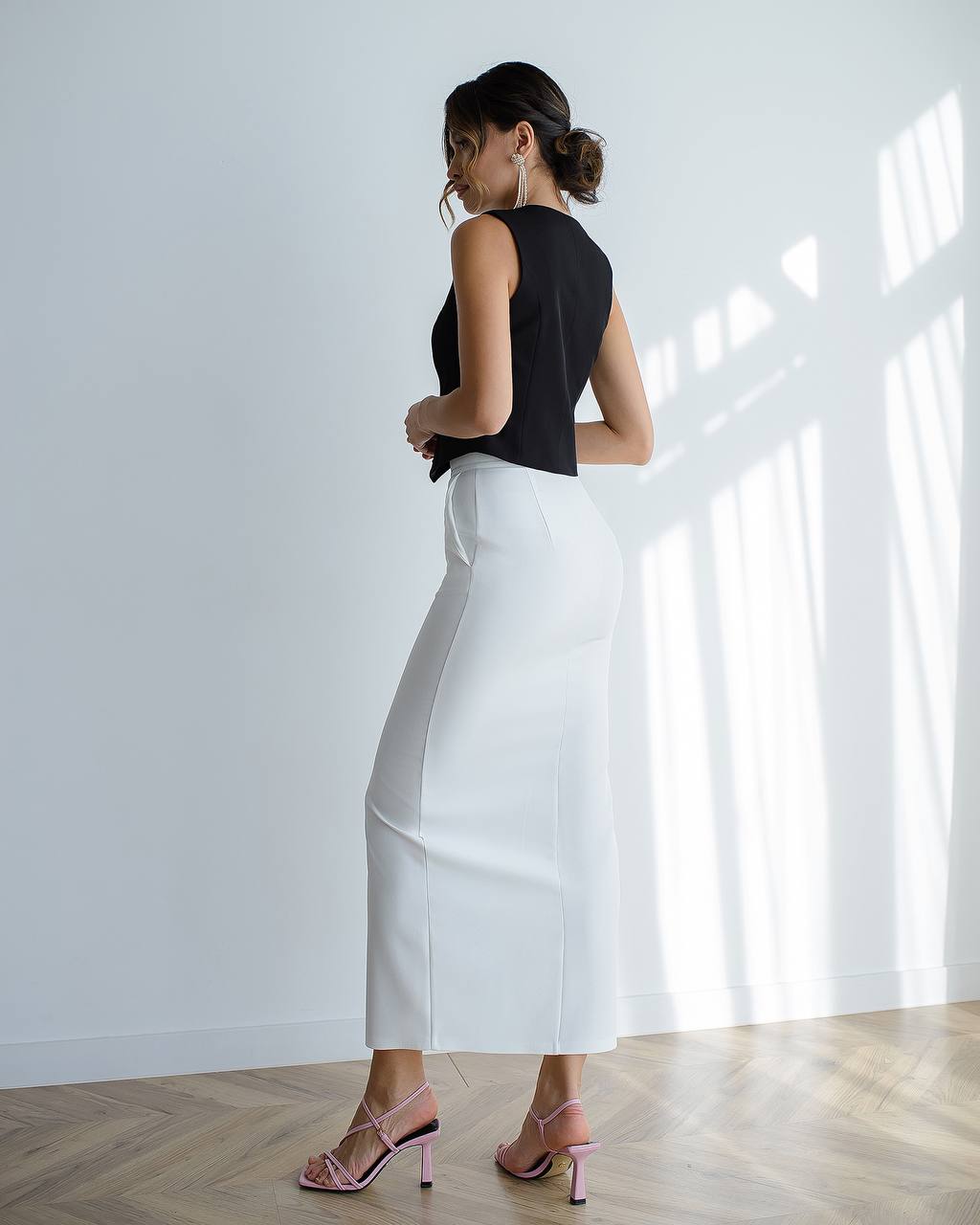 Woman wearing a black top and white skirt standing in a well-lit room with sunlight filtering through.