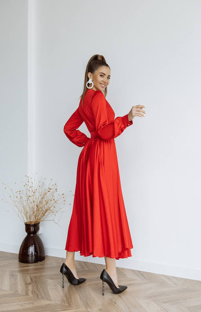 Woman in a red dress standing against a white wall with a vase of dried plants on a wooden floor.