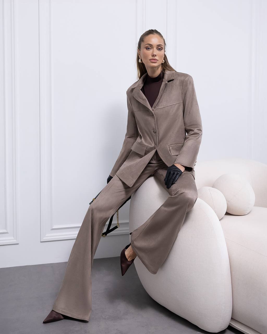 Woman in a beige suit sitting on a white chair against a white wall.