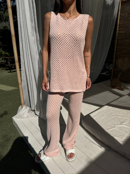 Person wearing a pink knitted outfit standing outdoors with light-colored curtains in the background.