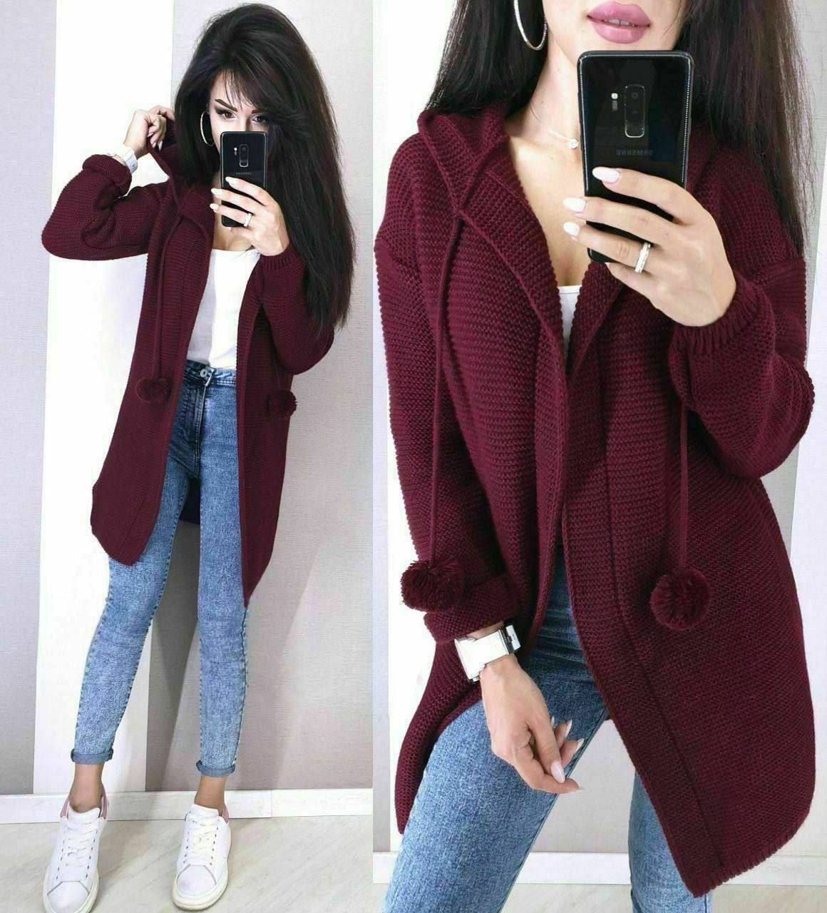 Woman wearing a burgundy cardigan with pom-poms, white shirt, and blue jeans, taking a mirror selfie.
