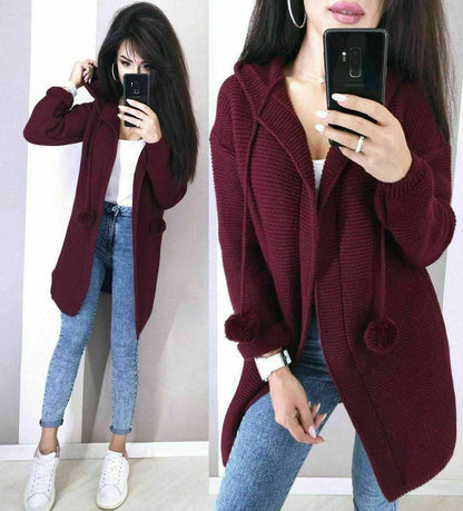 Woman wearing a burgundy cardigan with pom-poms, white shirt, and blue jeans, taking a mirror selfie.