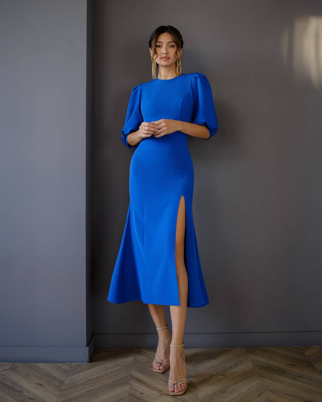 Woman wearing a blue dress with a slit standing against a gray wall.