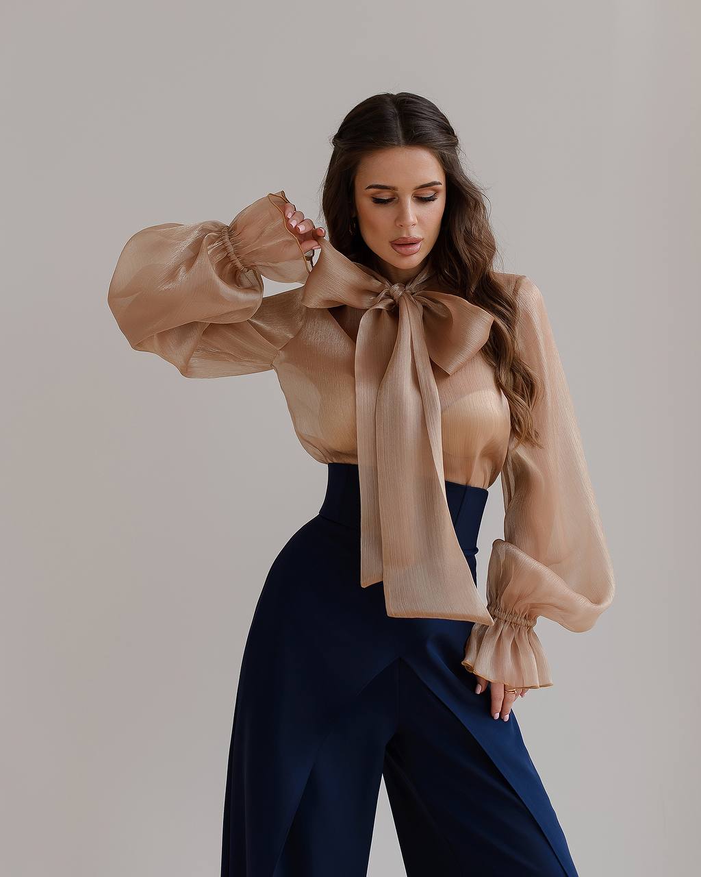 Woman wearing a beige blouse with a large bow and navy pants against a plain background