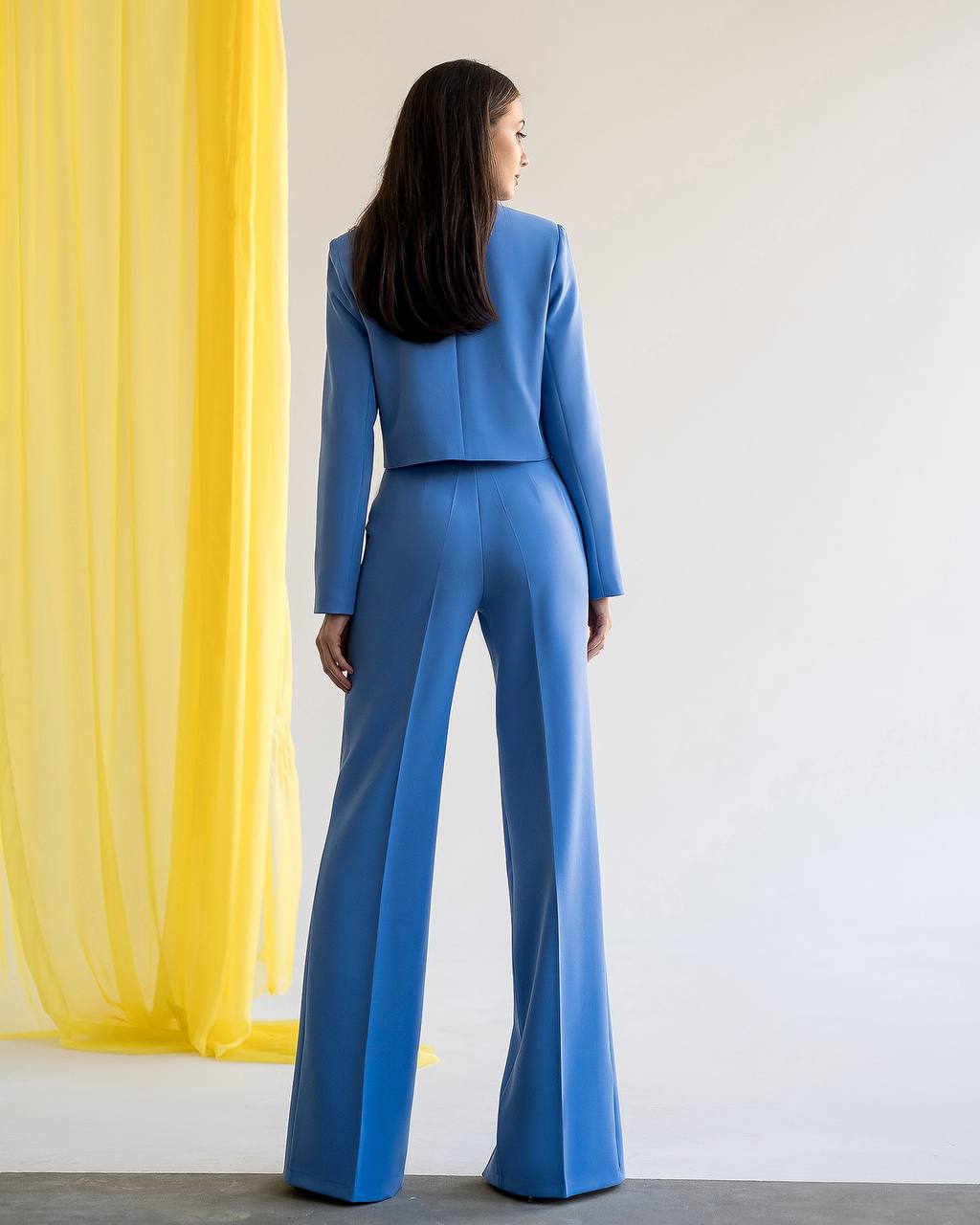 Woman wearing a blue suit standing in front of a yellow curtain and white wall.