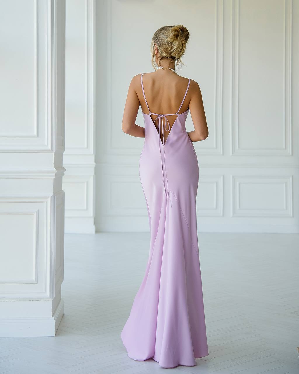 Woman wearing a long, light purple dress with thin straps in a white room.
