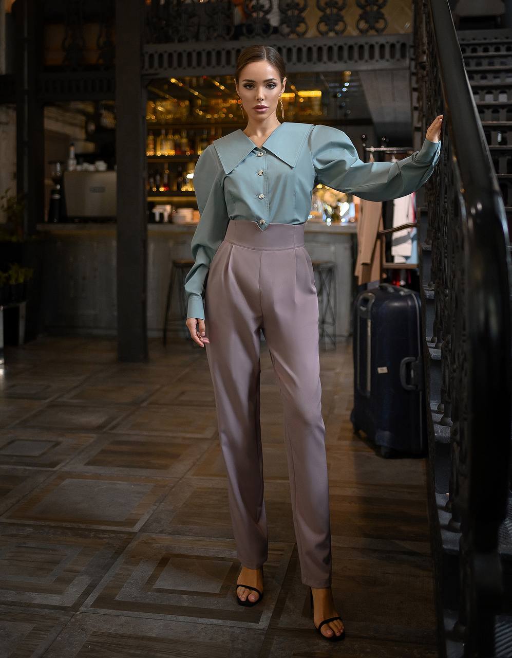 Woman in a light blue shirt and beige pants standing in a room with a bar in the background.