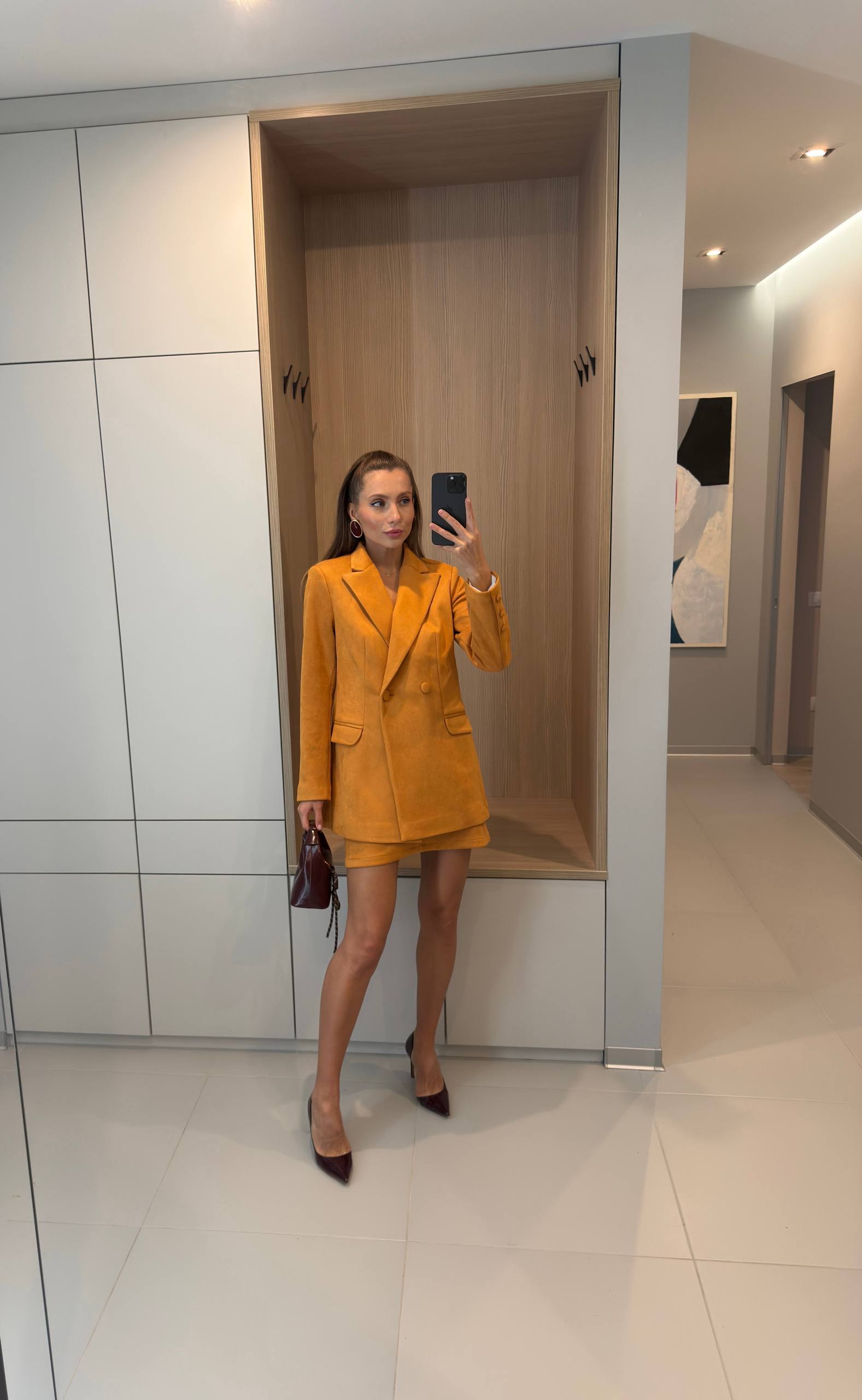 A woman takes a mirror selfie wearing a mustard yellow double-breasted blazer and matching mini skirt set, paired with dark brown pointed-toe heels, in a modern interior hallway with light-colored walls and built-in wooden storage compartments.