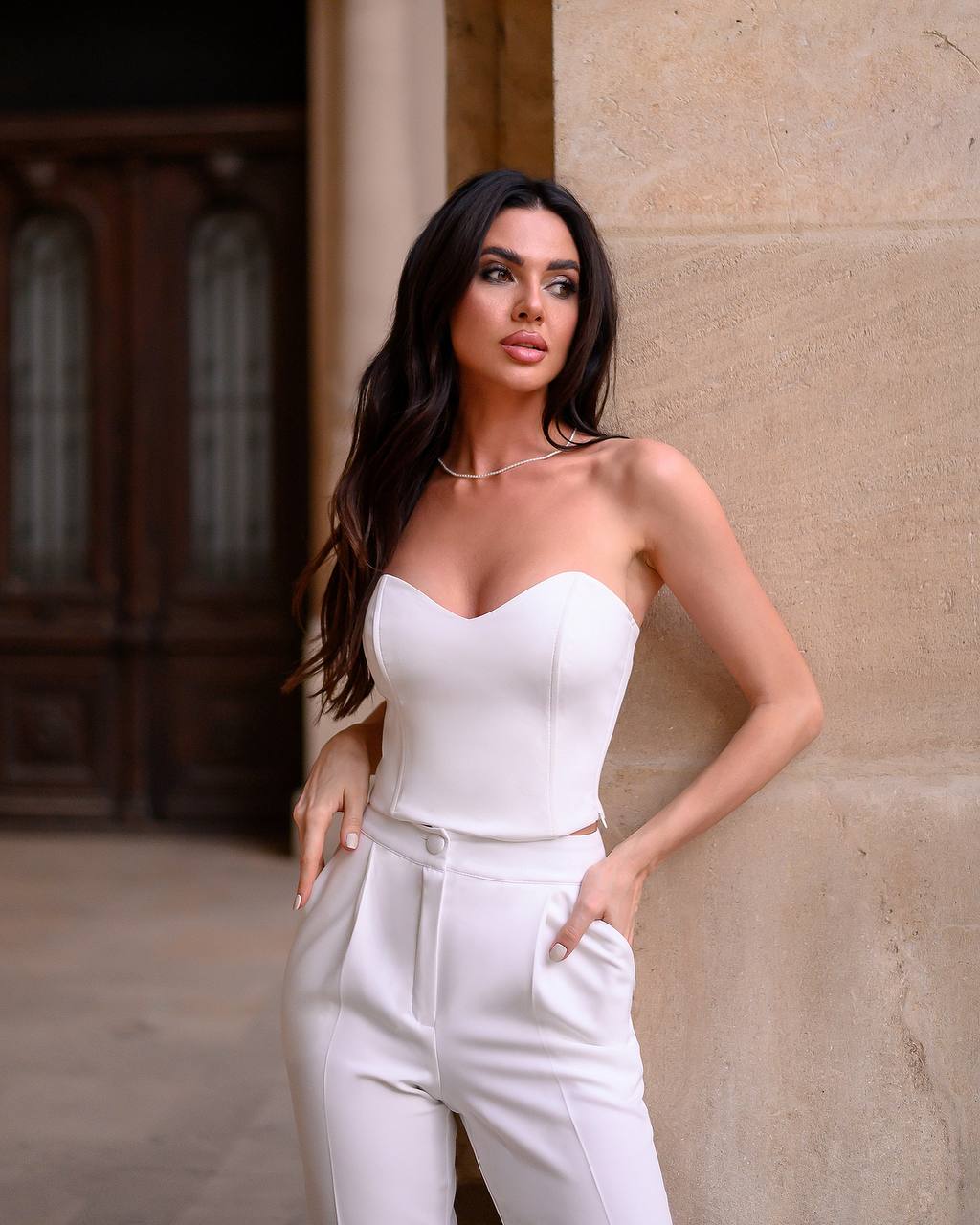 Woman in a white strapless top and high-waisted pants standing against a stone wall.
