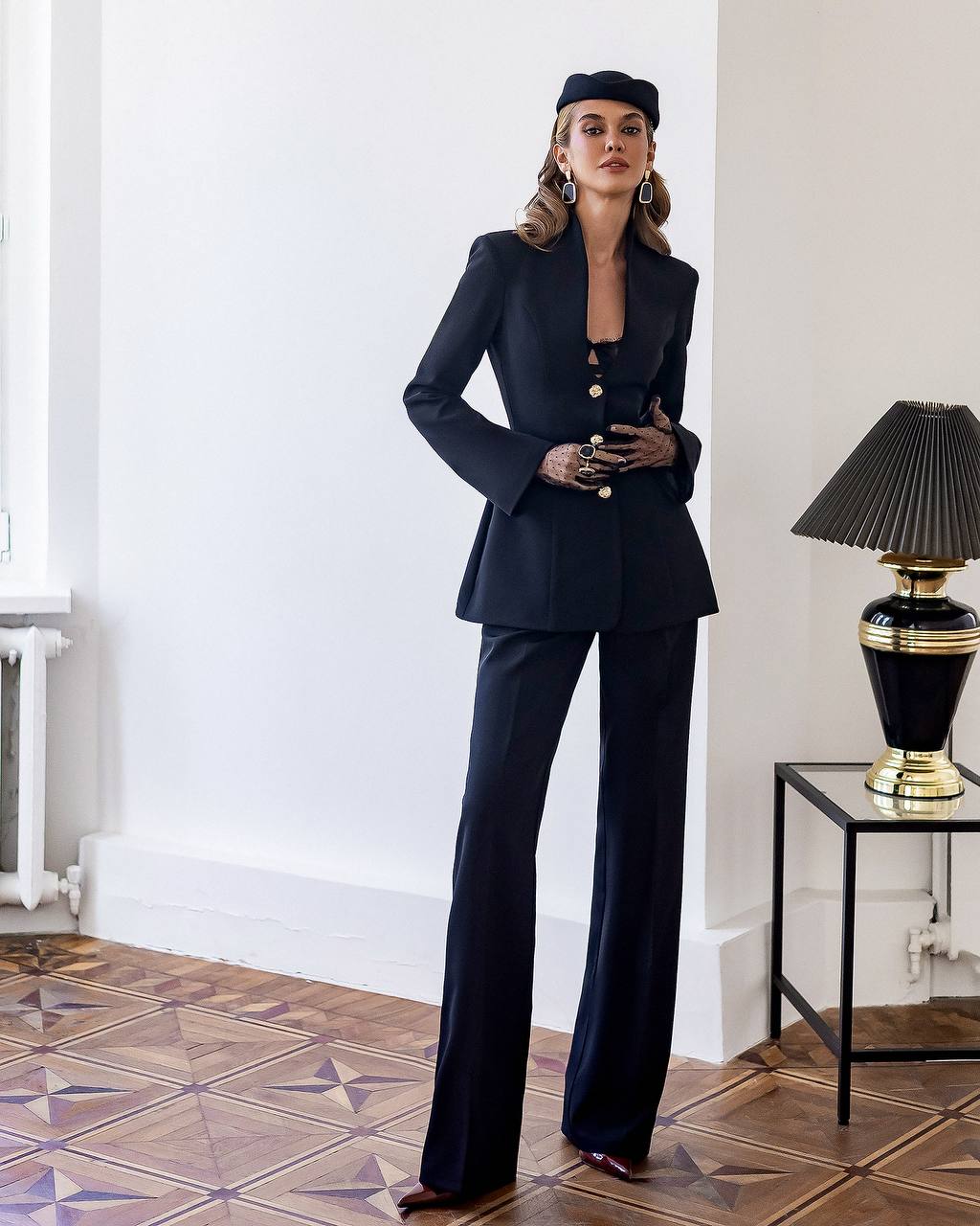 Woman in a black suit standing in a room with a lamp and table.