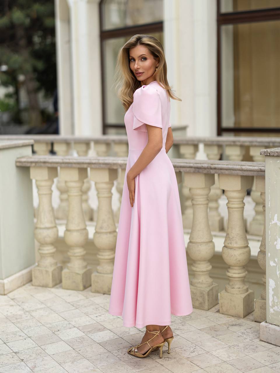 A woman wearing a light pink midi dress with short flutter sleeves and a flared skirt, standing on a stone terrace in front of a building with columns and windows.