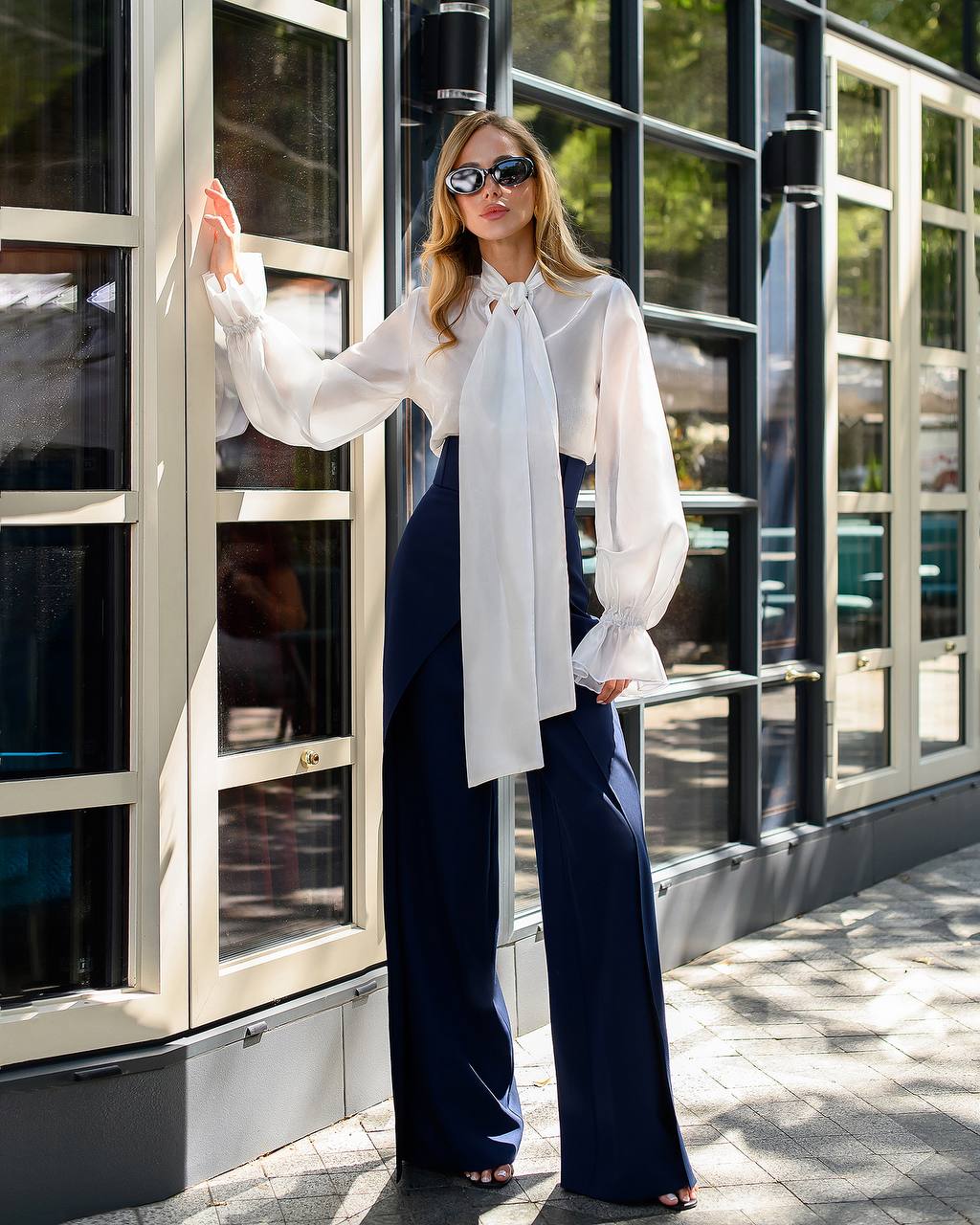 Woman wearing a white blouse with a large bow and navy pants standing in front of a building.