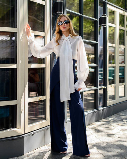 Woman wearing a white blouse with a large bow and navy pants standing in front of a building.