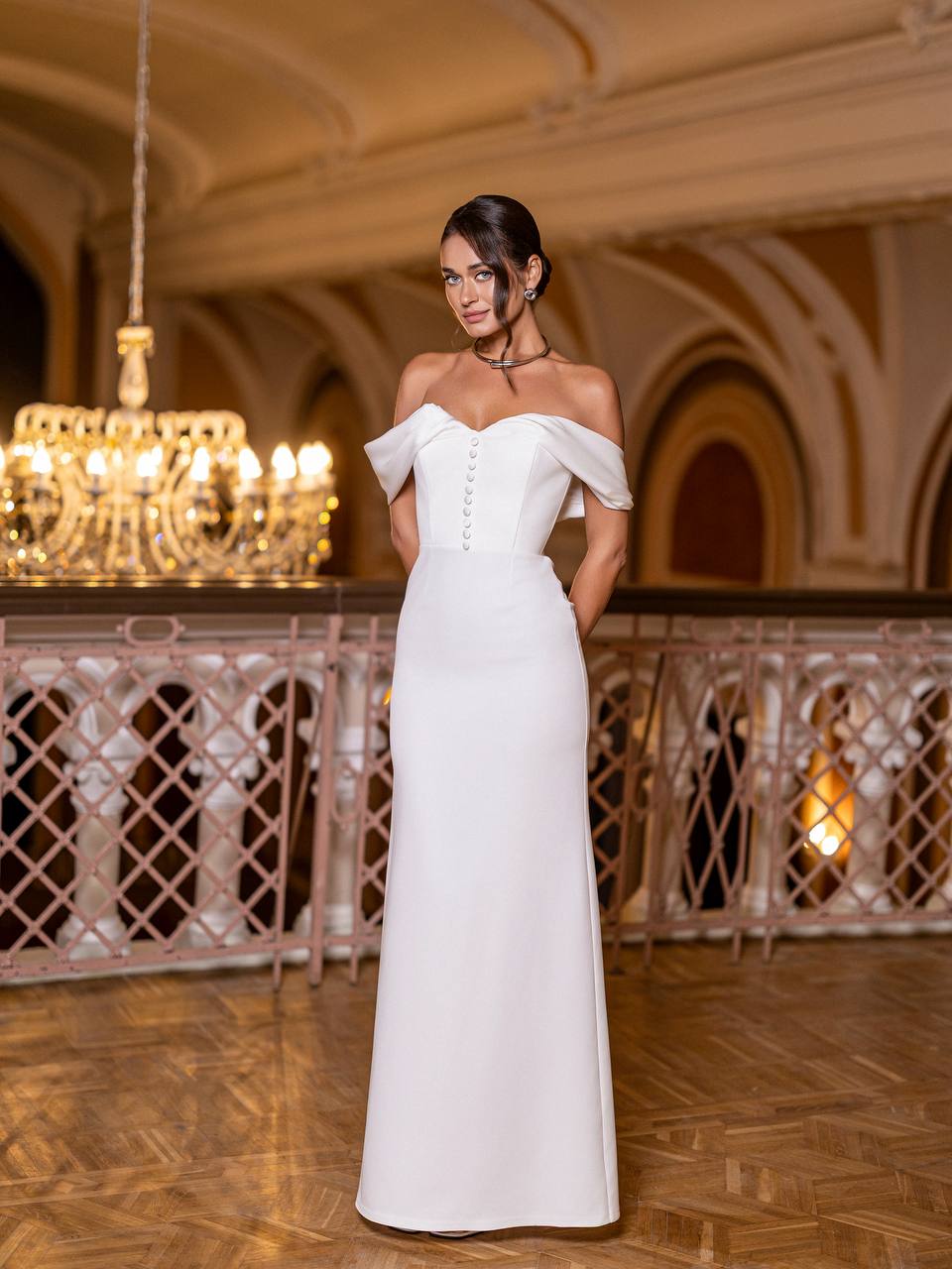 Woman in a white off-shoulder gown standing in an elegant indoor setting with chandeliers.