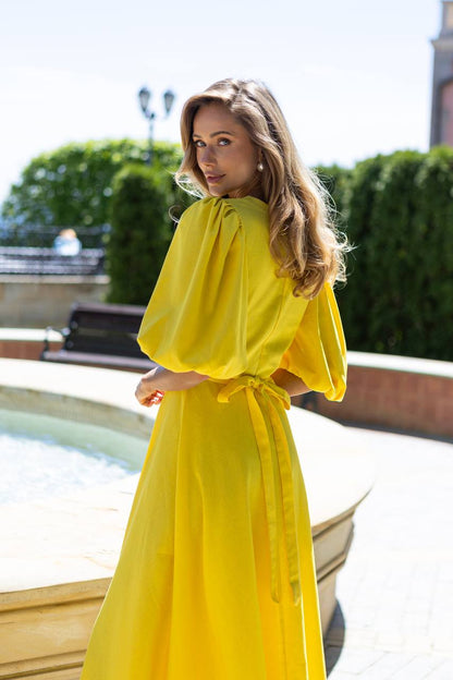 Woman in a yellow dress standing outdoors with greenery and a building in the background