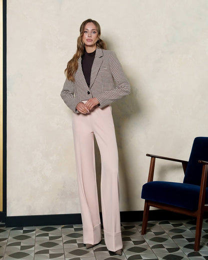 Woman wearing a checkered blazer and pink pants standing in a room with a chair and patterned floor.