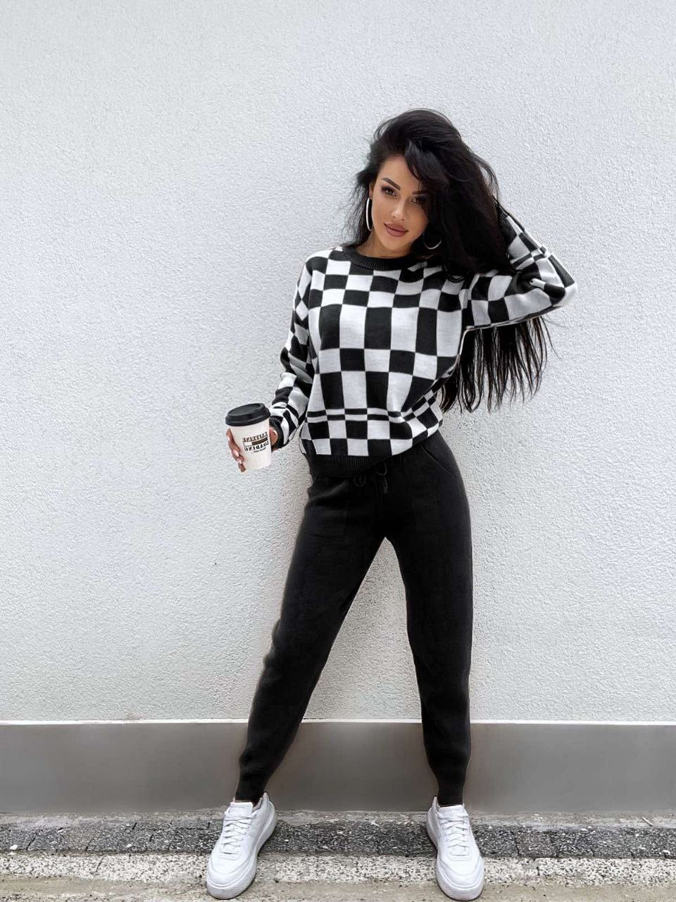 Woman wearing a black and white checkered sweater with black pants and white sneakers, holding a coffee cup against a light gray wall.